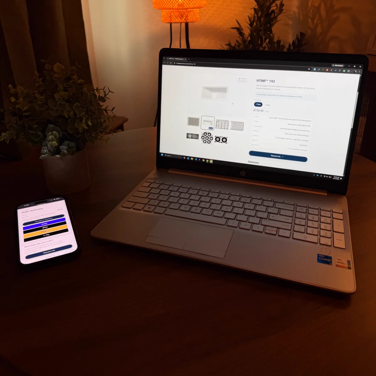 A laptop and phone on a wooden desk in a warm room, a store page open on the screen