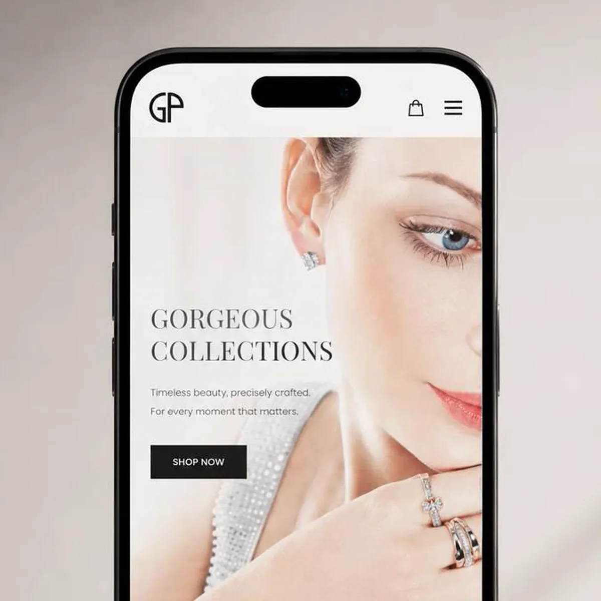 Smartphone standing on a pale stone surface against a soft beige background, showing a luxury jewelry brand's mobile home page: a close-up photo of a model wearing a diamond stud earring and two diamond rings, a large serif headline across the image, a two-line tagline, and a black Shop Now button.