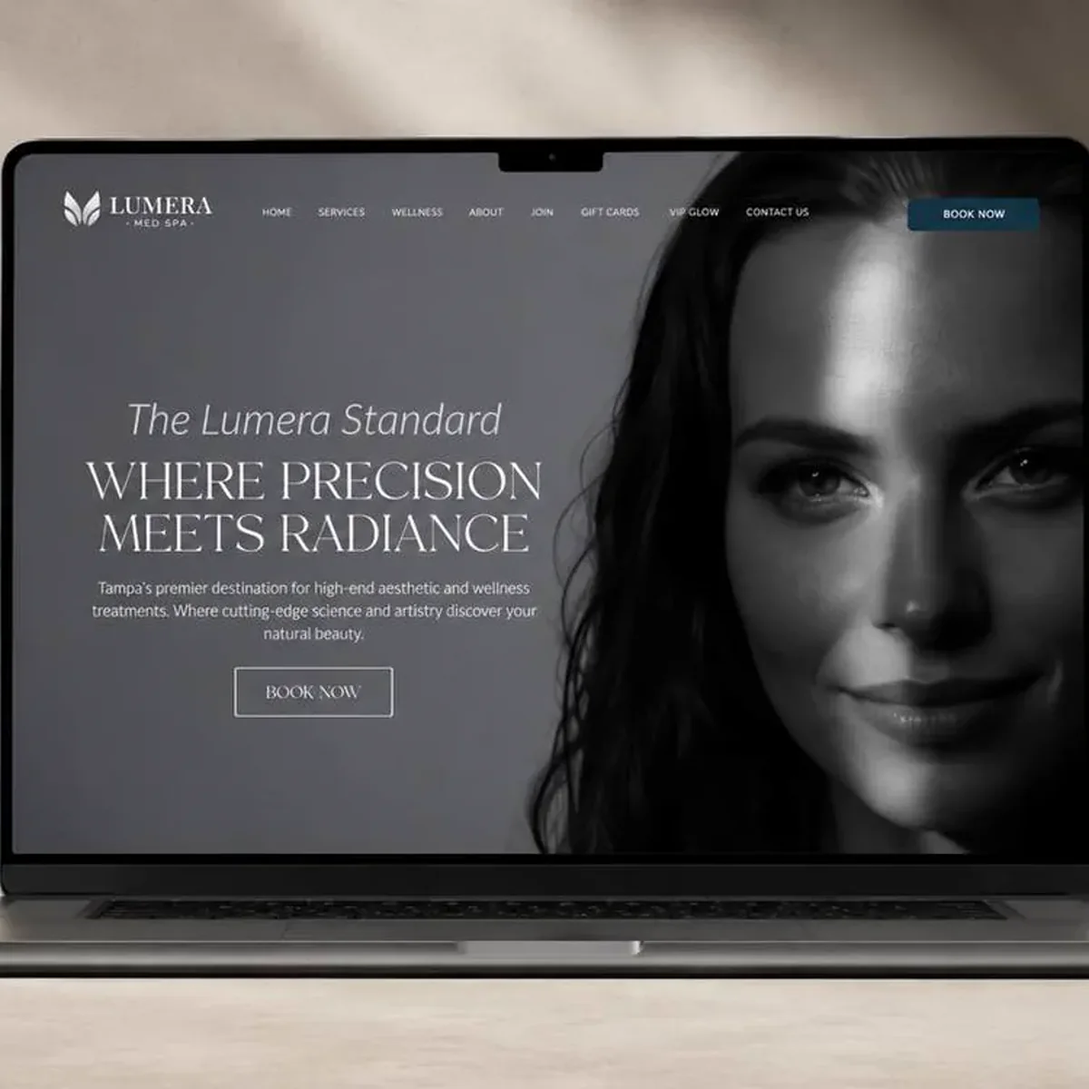A laptop on a pale desk displaying a med spa homepage. The charcoal hero places a black-and-white portrait of a smiling woman on the right and a tall serif headline, Where Precision Meets Radiance, on the left, above a short intro line and an outlined Book Now button, with a slim navigation bar and a teal Book Now button across the top.