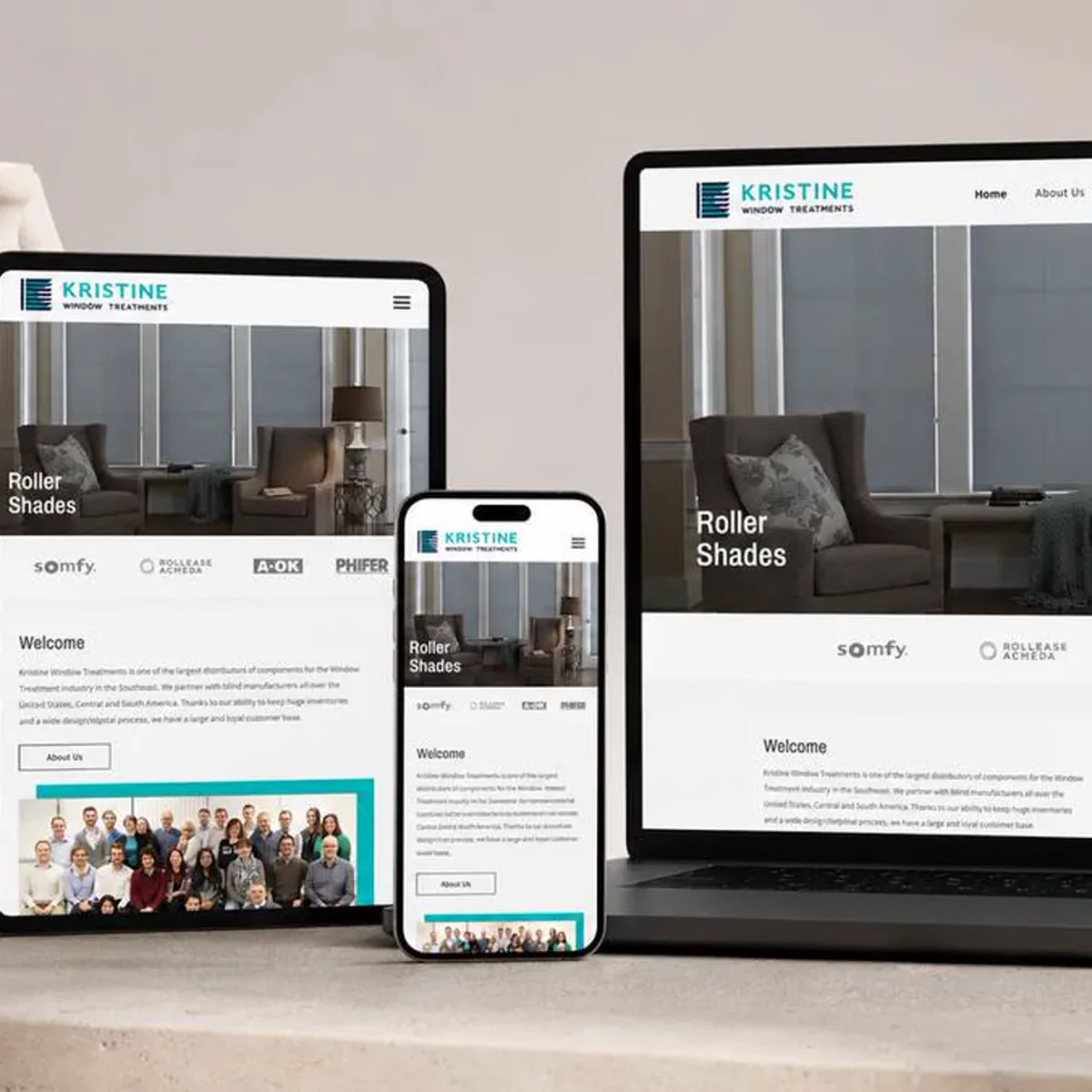 A tablet, a smartphone, and a laptop lined up on a stone ledge, each displaying the same window-treatment company homepage adapted to its own screen size, with a roller shade room photo, a row of partner logos, a welcome section, and a company team photo.