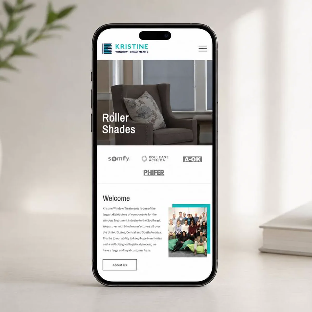 A smartphone standing upright on a pale desk beside a leafy plant and a stoneware cup, displaying the mobile homepage of a window treatments company: a logo and menu button at the top, a photo of a dark armchair beside a window fitted with a roller shade under the heading Roller Shades, a row of four supplier logos, and a Welcome section with a paragraph of text, a group photo of the staff, and an About Us button.